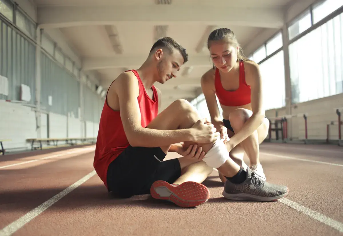 Two athletes in red sportswear sitting on an indoor track, one wrapping the other's leg with a white bandage