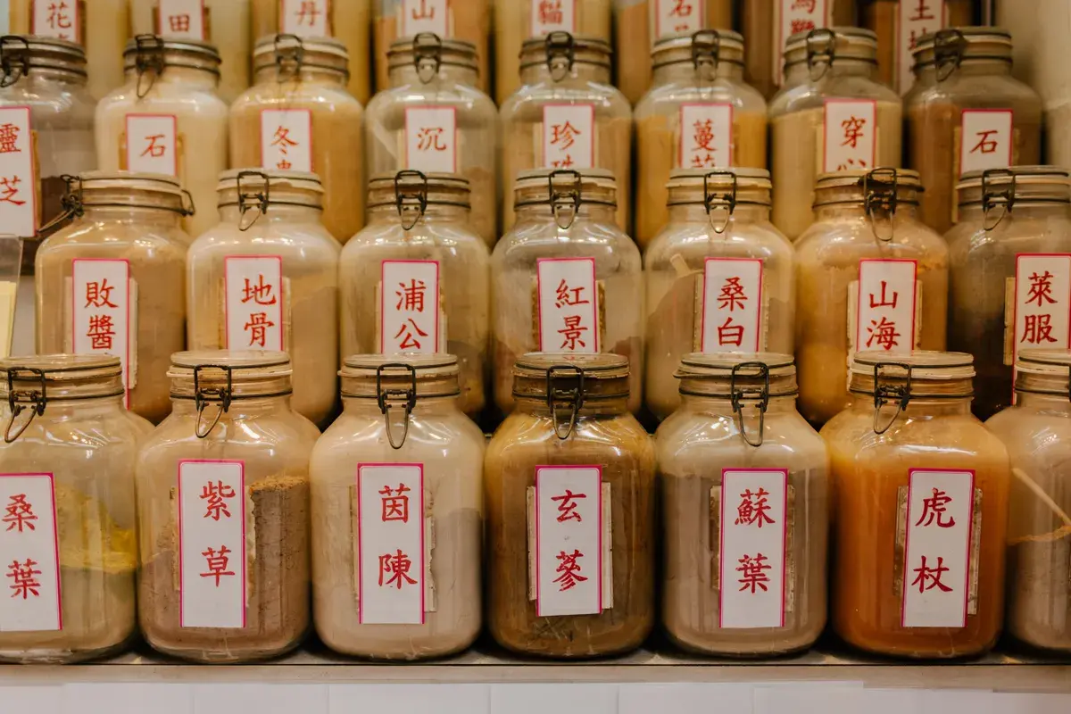 Glass jars with metal clasps containing dried herbs and plant materials, labeled with white tags bearing red Chinese