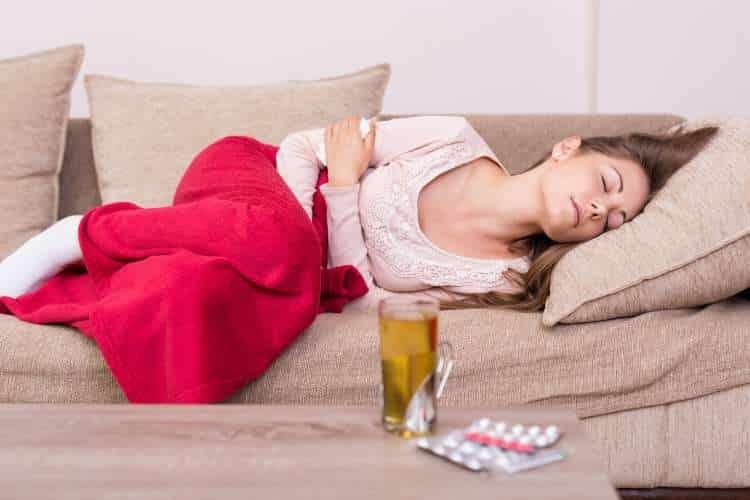 Woman resting on a sofa under a blanket with tea and tablets nearby